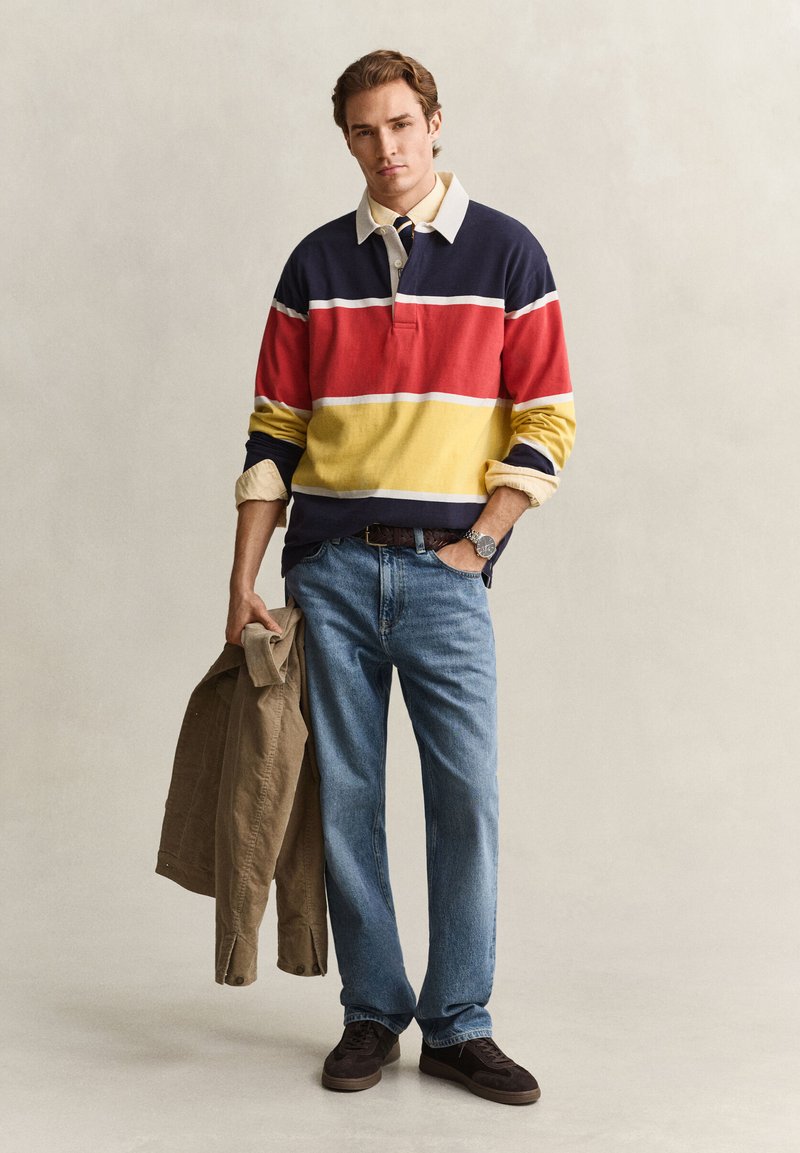 Mand iført en stribet langærmet skjorte, blå jeans og mørke sko, der holder en brun jakke med den ene hånd i lommen.