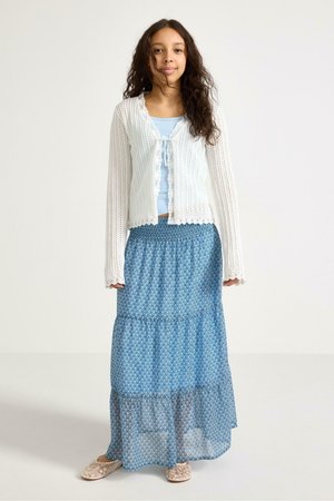 Nuori nainen seisoo valkoisessa neulotussa neuletakissa, vaaleansinisessä topissa, pitkässä sinisessä kuvioidussa hameessa ja vaaleanpunaisissa kukkakuvioisissa tohveleissa yksiväristä taustaa vasten.