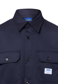 Camisa de manga corta de un oscuro color navy con cuello, dos bolsillos delanteros, botones negros y una etiqueta de marca en el bolsillo izquierdo. Tejido texturizado.