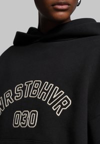 Black sweatshirt featuring a hood, with textured embroidery reading "NRSTBHR" and "030" in a contrasting cream color.