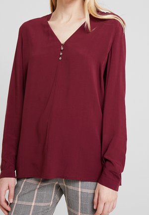 Frau trägt eine langärmelige burgunderrote Bluse mit V-Ausschnitt und vier Knöpfen, kombiniert mit schwarz-weiß karierten Hosen mit rosa Streifen.