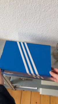 Blue shoe box with three white zigzag stripes on the lid. The box is rectangular with a smooth surface and a logo on one side.