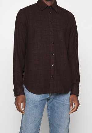 Mørkebrunt ternet langærmet skjorte med knapper, båret med lyseblå jeans på en stående person mod en ensfarvet baggrund.