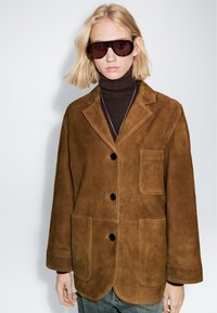 Blazer in suede marrone con vestibilità rilassata, dotato di colletto a revers, due tasche frontali e bottoni neri. Indossato sopra un dolcevita scuro.