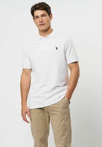 Młody mężczyzna w białej koszulce polo i beżowych spodniach, stojący z jedną ręką w kieszeni na gładkim jasnym tle.