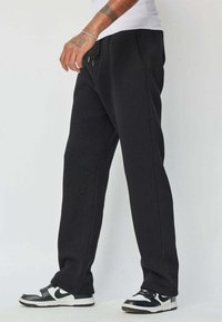 Pantalons de survêtement noirs en coton, avec une coupe décontractée, une taille élastique avec cordon de serrage et des poches latérales. Associés à des baskets noires et blanches.