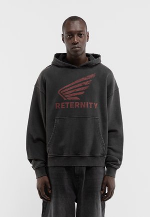 Mann trägt schwarzen Hoodie mit rotem Flügellogo und dem Wort "RETERNITY", steht vor einem schlichten hellen Hintergrund.