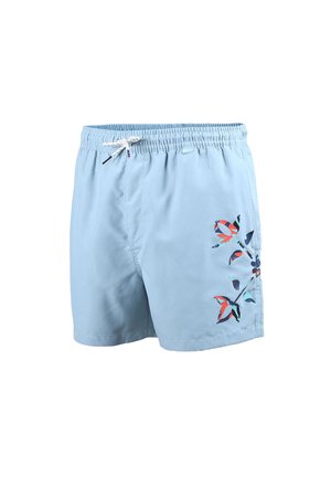 Jeune homme debout pieds nus portant un short de bain bleu clair avec lien de serrage et motif floral sur la jambe droite, fond neutre.