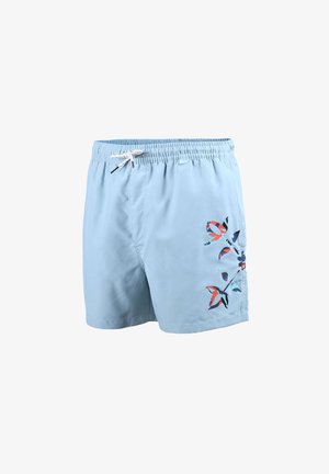 Jeune homme debout pieds nus portant un short de bain bleu clair avec lien de serrage et motif floral sur la jambe droite, fond neutre.