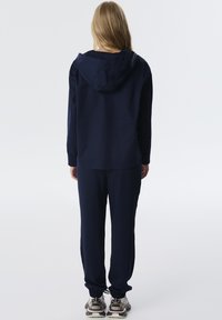 Chándal navy con una sudadera con capucha, puños acanalados y pantalones elásticos. El modelo lleva zapatillas acolchadas de color claro.