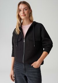 Felpa nera con cerniera in morbido tessuto, con due tasche frontali, coulisse regolabili sul cappuccio, indossata con una maglietta rosa chiaro.