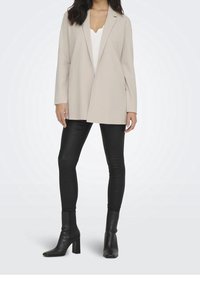 Béžový oversized blejzr s jedním knoflíkem, dlouhými rukávy a otevřeným předním dílem, v kombinaci s černými koženými legínami a botami na podpatku.
