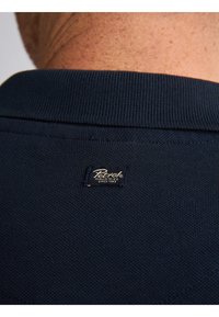 Navy blue polo-paita, jossa on teksturoitu neulos. Varustettu ribbikauluksella ja brändätystä etiketistä niskassa.