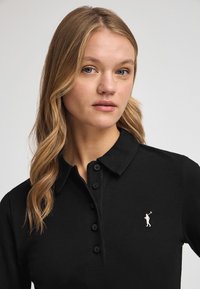Camisa polo negra de tejido texturizado, con un collar clásico y seis botones, que presenta un pequeño logotipo bordado de golfista en el lado izquierdo.