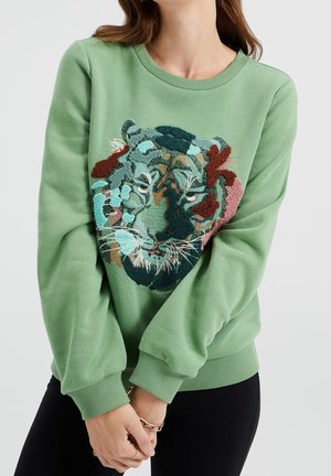 Femme portant un sweat-shirt vert avec un motif texturé, brodé multicolore représentant un visage de tigre, mains jointes devant, sur fond uni.