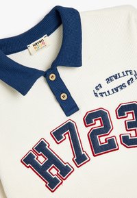 Camiseta tipo polo para niño pequeño con un cuerpo blanco acanalado, cuello azul marino, dos botones de madera y un diseño bordado en rojo que presenta "H723".