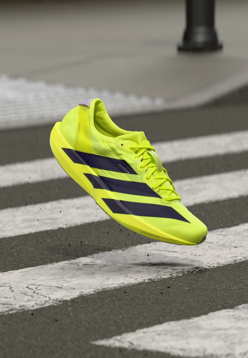 Jasnożółty but sportowy z ciemnofioletowymi paskami. Lekka konstrukcja, gładka tekstura i niska sylwetka, przedstawione w powietrzu nad przejściem dla pieszych.