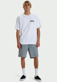 Jovem com cabelo loiro a usar uma t-shirt gráfica branca, calções cinzentos, meias brancas e sapatos pretos sem atacadores, a estação contra um fundo simples.