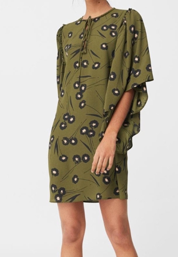 Femme portant une robe courte fleurie vert olive avec des manches larges et un détail noué au niveau de l'encolure, debout devant un fond uni.