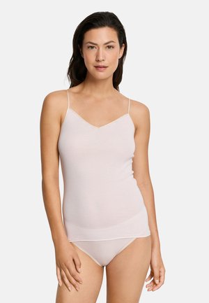 Kvinde med mørkt hår iført lysebeige camisole og matchende undertøj, stående foran en ensfarvet hvid baggrund, vendt fremad.
