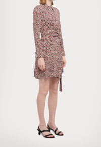 Robe à manches longues avec boutons, présentant un motif géométrique rouge et noir, ceinture à la taille et tissu léger ; portée avec des sandales à talons noires.