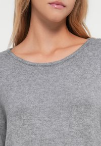 Pull gris clair en tricot doux. Il présente un col rond et des bords côtes, mettant en valeur une texture lisse et un design simple.