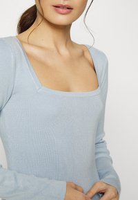 Haut long-sleeve côtelé bleu clair avec un décolleté carré et une coupe ajustée. Texture douce avec un éclat subtil, mettant en valeur le décolleté et les manches.