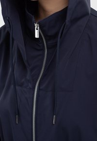 Navy blue jacket with a high collar and adjustable drawstrings, featuring a silver zipper and smooth, water-resistant fabric.