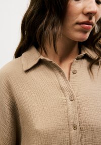 Beige textured shirt with a collared neck, button-down front, and a crinkled fabric pattern. Close-up view of the neckline and shoulder.