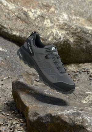 Chaussure de randonnée Columbia gris foncé avec semelle texturée qui semble flotter au-dessus d'un terrain rocailleux accidenté.