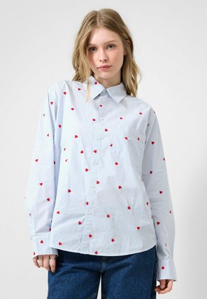 Jonge vrouw draagt een lichtblauw gestreept overhemd met kleine rode hartjes en een donkere spijkerbroek, staand tegen een effen achtergrond.