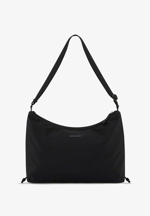 Personne portant un pull sans manches gris sur une chemise blanche, un pantalon noir à jambes larges, des chaussures noires pointues et portant un grand sac à épaule noir.