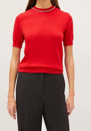 Vrouw met een rood sweater met korte mouwen en een parelkettingdetail bij de kraag, gecombineerd met zwarte hoge taillebroek.