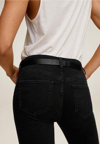 Henkilö, joka pitää yllään valkoista olkatoppaamattomaa paitaa, joka on työntänyt tiukkoihin mustiin farkkuihin mustalla vyöllä, kädet lantiolla, kasvot poispäin.