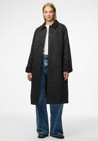 Cappotto nero lungo con collo a revers, bottoni frontali e tasche laterali; abbinato a jeans a gamba larga, blu sbiadito. Tessuto liscio e opaco.