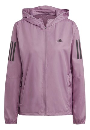 Personne portant une veste à capuche zippée Adidas couleur violet clair, les mains dans les poches et un legging de sport assorti.
