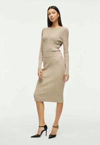 Robe en tricot beige avec une texture côtelée, des manches longues et une silhouette ajustée, associée à des chaussures à talons hauts noires.