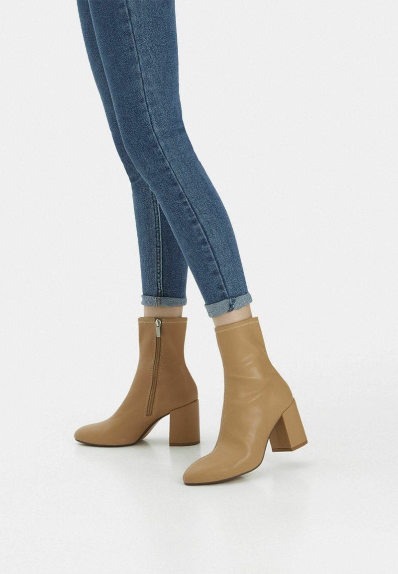 Botines marrones claros con punta afilada y tacón cuadrado. Hechos de piel suave, con una cremallera lateral y acentos cosidos. Combinados con jeans remangados.