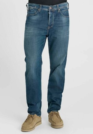 Blauwe jeans met rechte pijpen gedragen met beige suède veterschoenen bij een persoon die tegen een effen achtergrond staat.