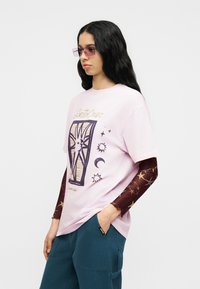 Γυναίκα με μακριά μαύρα μαλλιά που φοράει ροζ γραφικό t-shirt, μακρυμάνικο με μοτίβα μπορντό, τιρκουάζ παντελόνι και ροζ ορθογώνια γυαλιά ηλίου, στέκεται με το χέρι στην τσέπη.