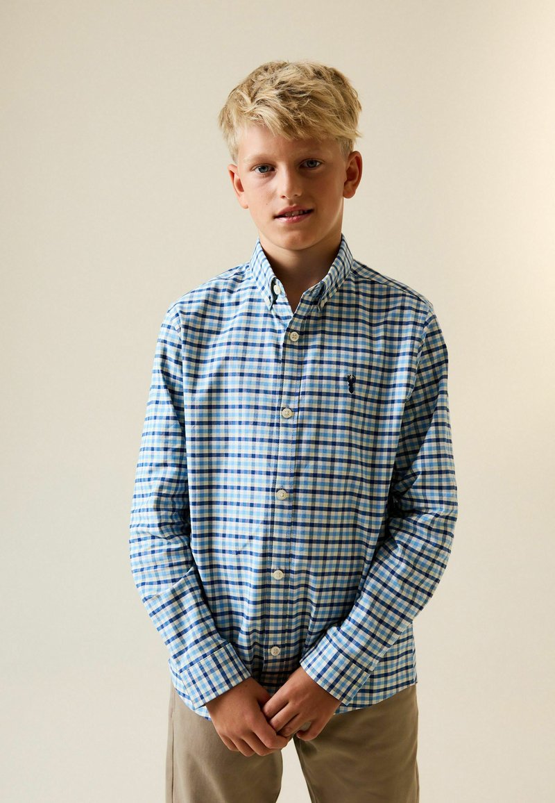 Blau-weiß kariertes Hemd mit Knopfleiste und offenem Kragen. Lange Ärmel, eine Brusttasche und Knopfverschluss. Helle, beige Hose.