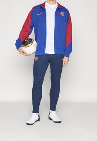 Nike Barcelona trackjacka i blått och rött, kombinerad med marinblå ribbade byxor. En person med ett mönstrat fotboll håller vita sportskor.