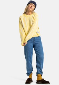 Geel gebreid sweater met witte strepen, ronde hals, losse pasvorm. Gecombineerd met blauwe jeans, donkerbruine schoenen en mosterdkleurige sokken.