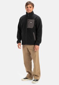 Mustmustest musta kõrge kaelusega jakk, millel on tõmblukuga rinnakott ja küljetaskud; omab pehmet tekstuuri ja minimaalseid halli aktsente.