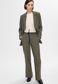 Olive rohelise tooniga kohandatud ülikond, millel on struktureeritud bleiser ja laiad püksid. Omadused: pehme materjal, allpool kreemjas topp ja must nöör.