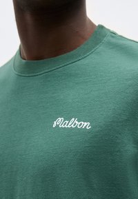 Gros plan sur une personne portant un t-shirt vert à col rond avec le texte blanc "Malbon" brodé sur la poitrine.