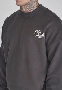 Sudadera gris oscuro de tejido suave. Presenta un cuello redondo y un logo blanco en el lado izquierdo del pecho. Textura suave y corte relajado.
