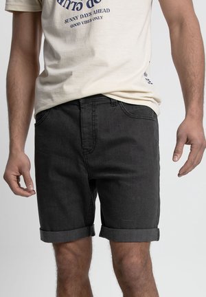Shorts en denim noir avec ourlet roulé, dotés de poches avant et d'une texture lisse. Portés avec un t-shirt graphique de couleur crème.