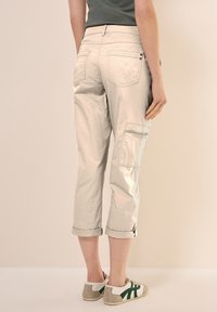 Lichtbeige cropped cargo broek met opgerolde manchetten, zij- en achterzakken, en een gladde textuur, gecombineerd met witte en groene sneakers.