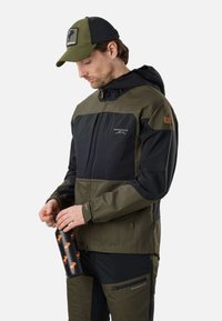Homme en veste d'extérieur verte et noire et casquette tenant une bouteille noire avec des motifs d'élan orange.